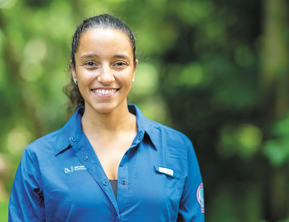 Portrait outdoor photograph close-up view of Jennifer Barros, Ph.D. smiling and standing in a blue colored Bat Conservation International branded button-up dress shirt