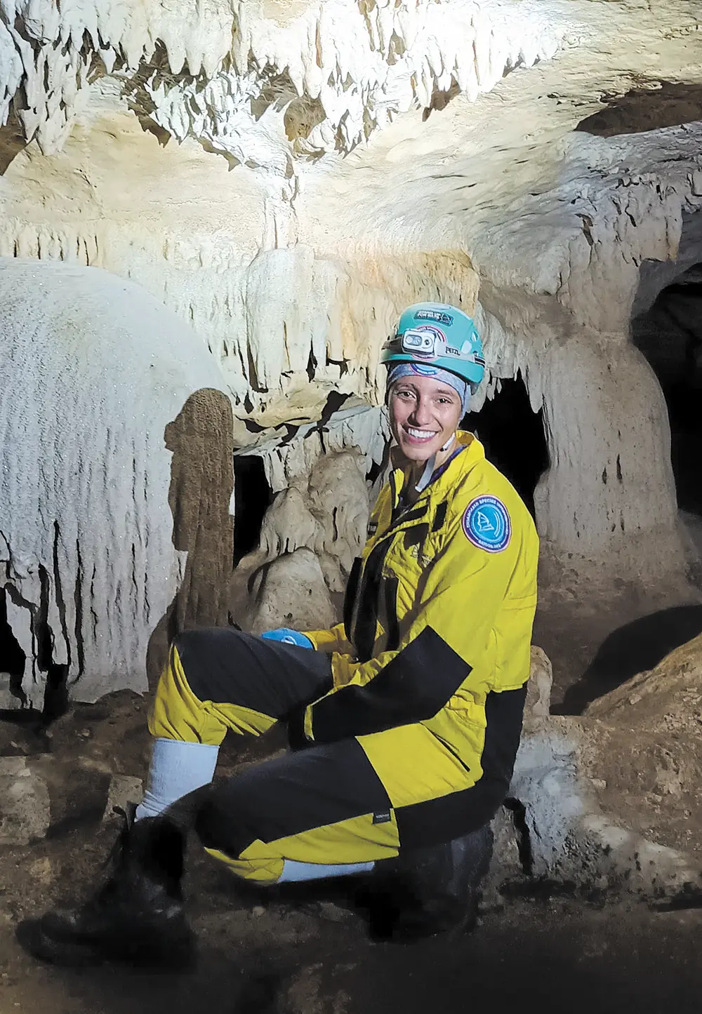 Portrait indoor photograph close-up view of Jennifer Barros, Ph.D. smiling and seated down on a cavern ground area in a yellow/black/blue colored Bat Conservation International jumpsuit outfit and a light turquoise hardhat equipped with a flashlight on it as she explores a cave in Brazil