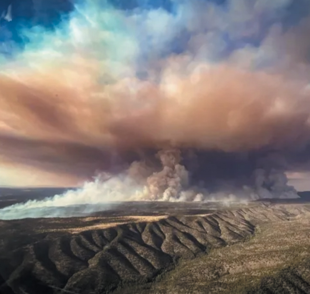 Aerial view of a large wildfire with thick smoke rising over rugged terrain and hills.