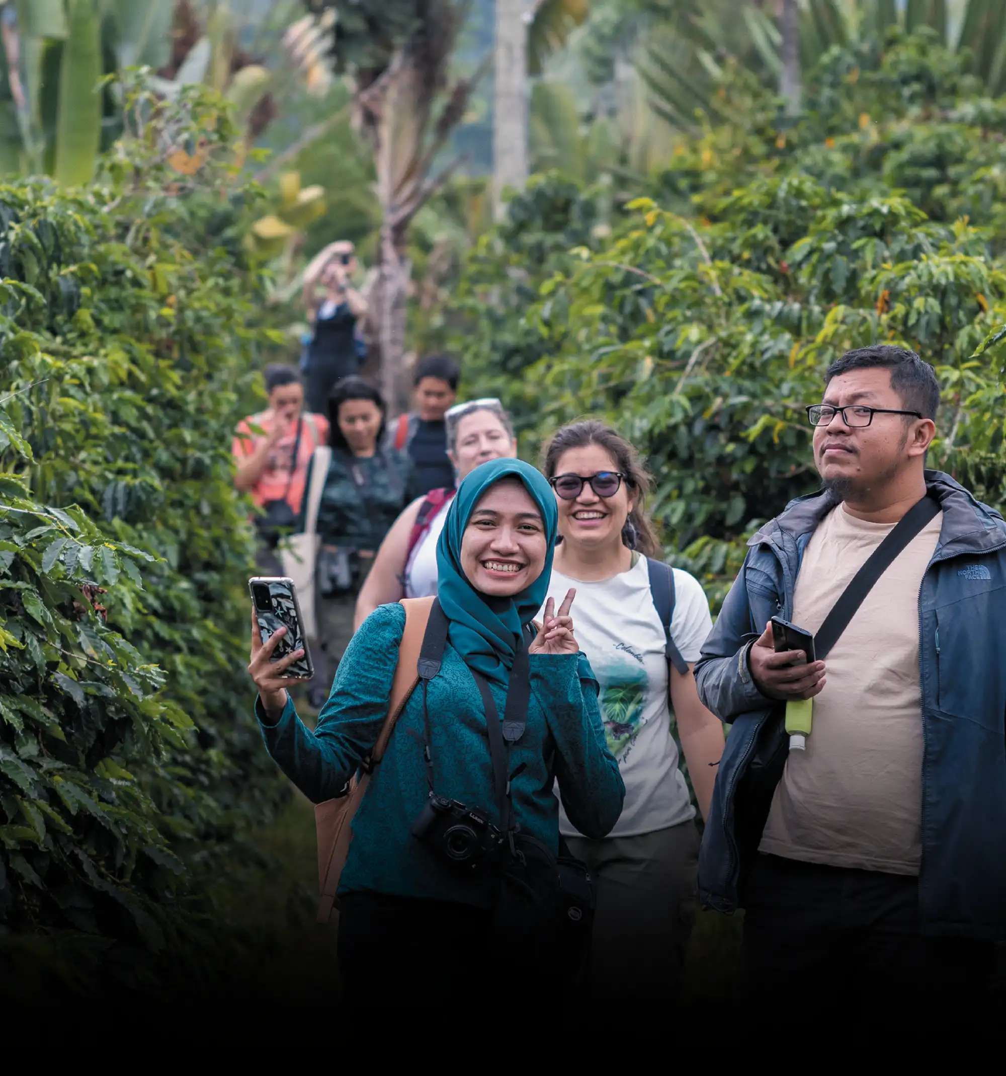 A group of people walking through a lush plantation, with a woman in a teal hijab making a peace sign in the foreground.