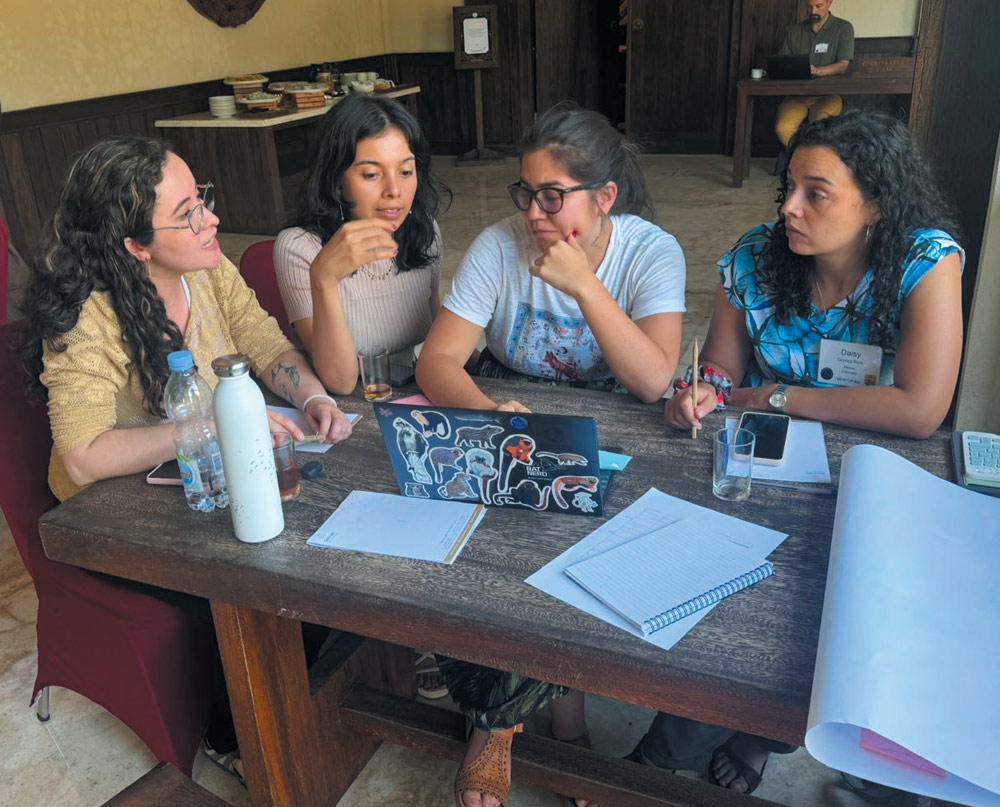 Four women engaged in discussion at a wooden table with a laptop and notebooks.