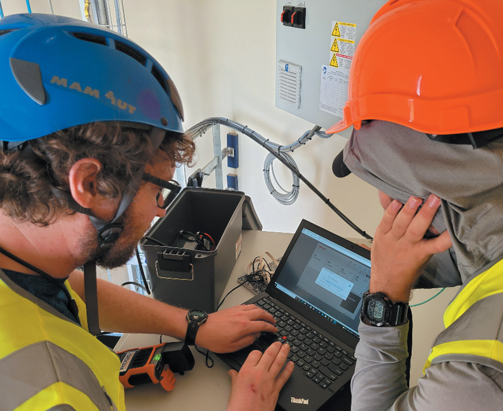 Two individuals with safety gear work on a laptop in an industrial setting.