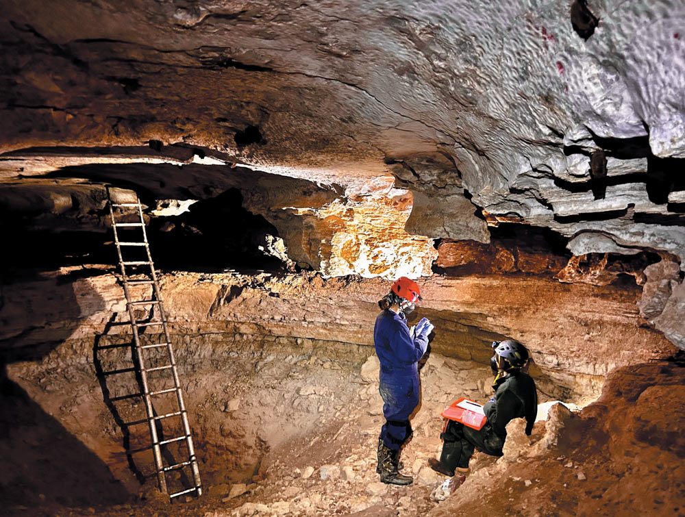 Two people in protective gear inside a cave, with a ladder and rugged rocky surfaces.