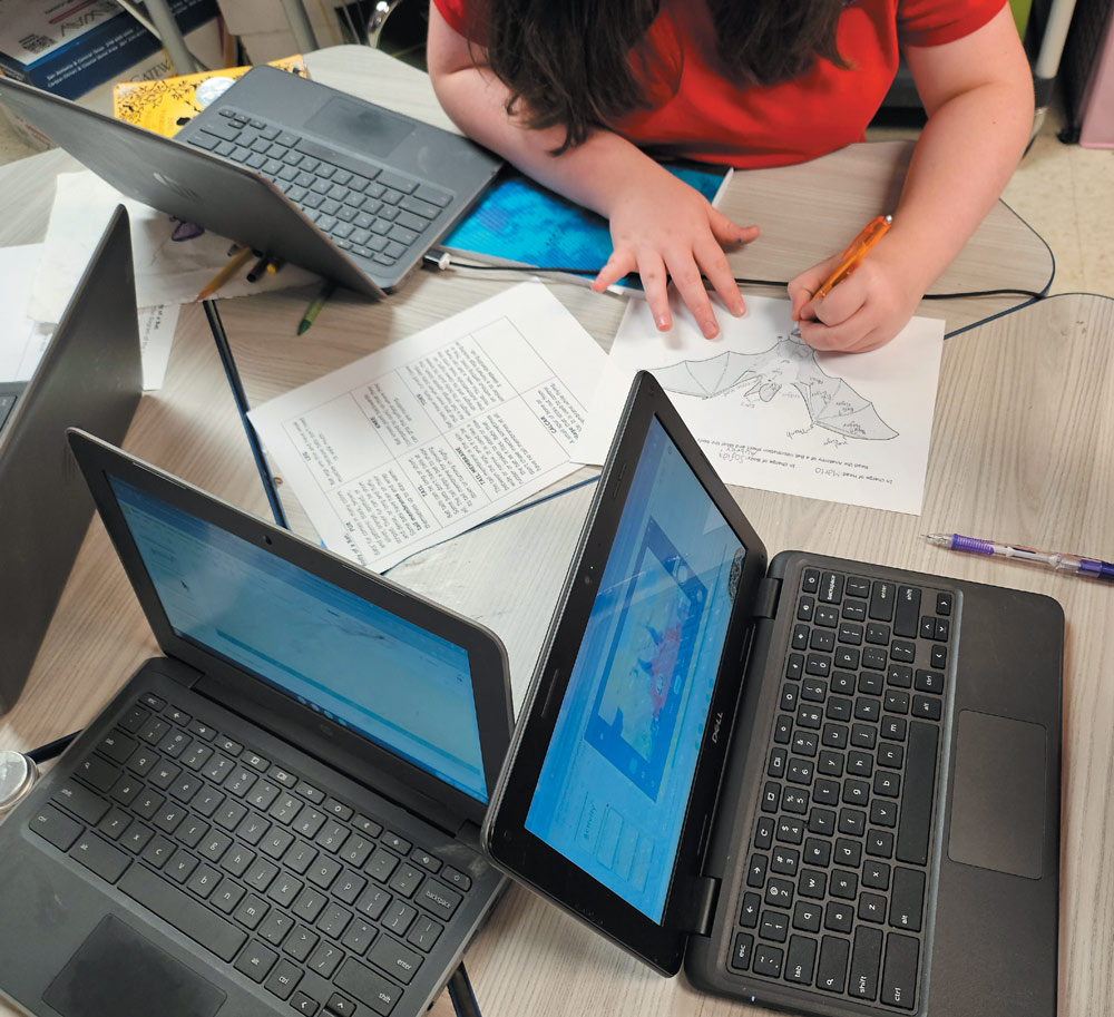 A person writing on paper surrounded by laptops and documents on a table.