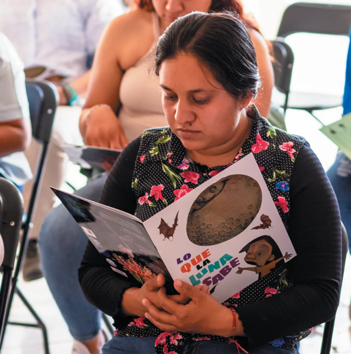 A seated woman reading a colorful educational booklet about bats during an indoor community event.
