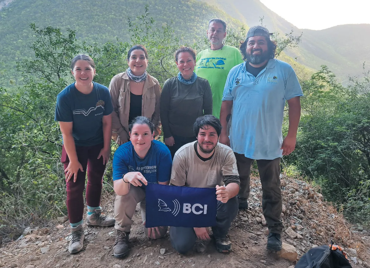 A group of seven people posing together on a dirt path with lush green hills in the background, two of them kneeling and holding a "BCI" flag.