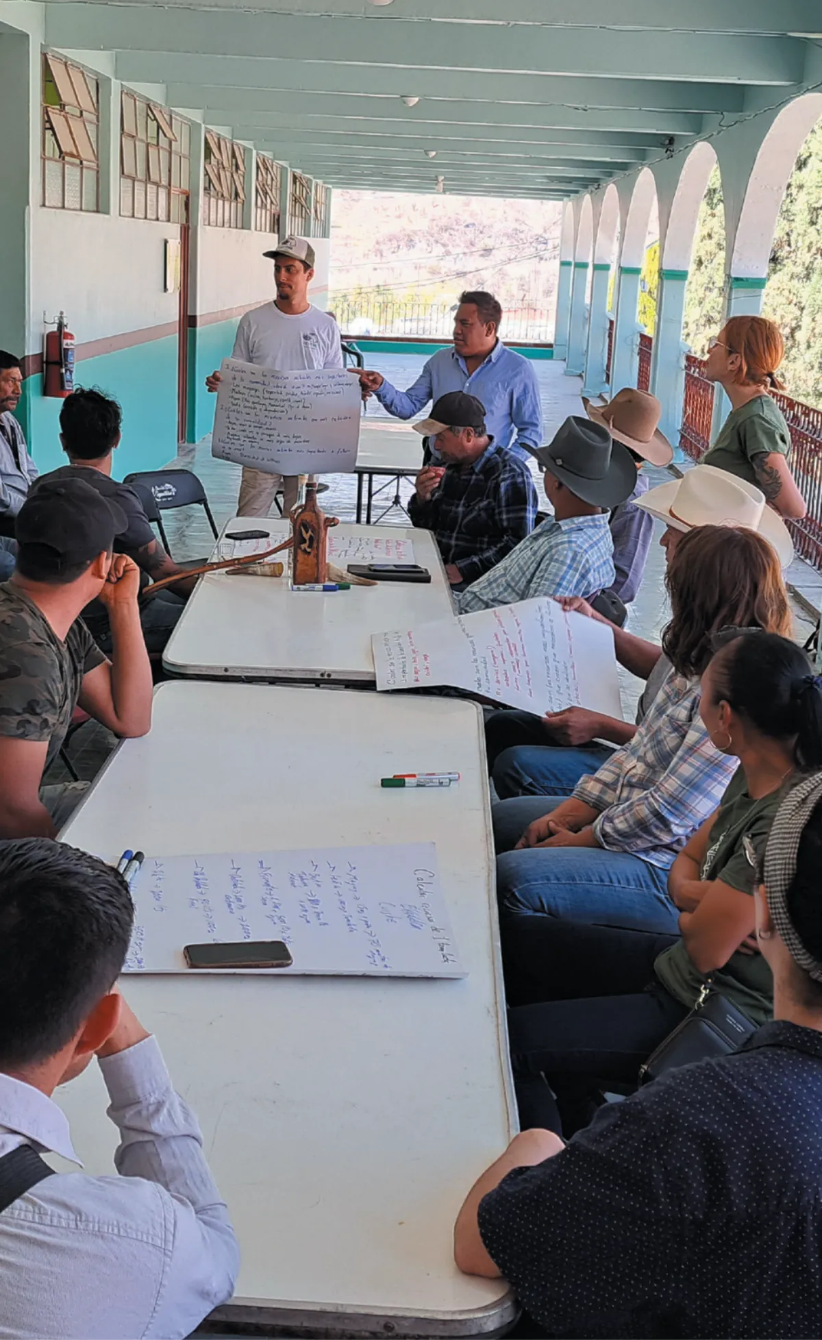 A group of people seated around tables, listening to a man holding a large sheet of paper with handwritten notes during a workshop in an outdoor corridor.