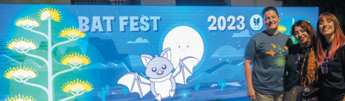 Three people posing in front of a vibrant "Bat Fest 2023" display featuring a cartoon bat and desert-themed artwork under nighttime lighting.