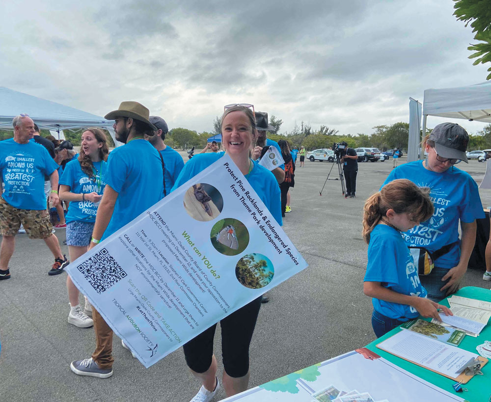 People at an outdoor event, one holding a "Protect Pine Rocklands" poster, nearby attendees wearing blue t-shirts.