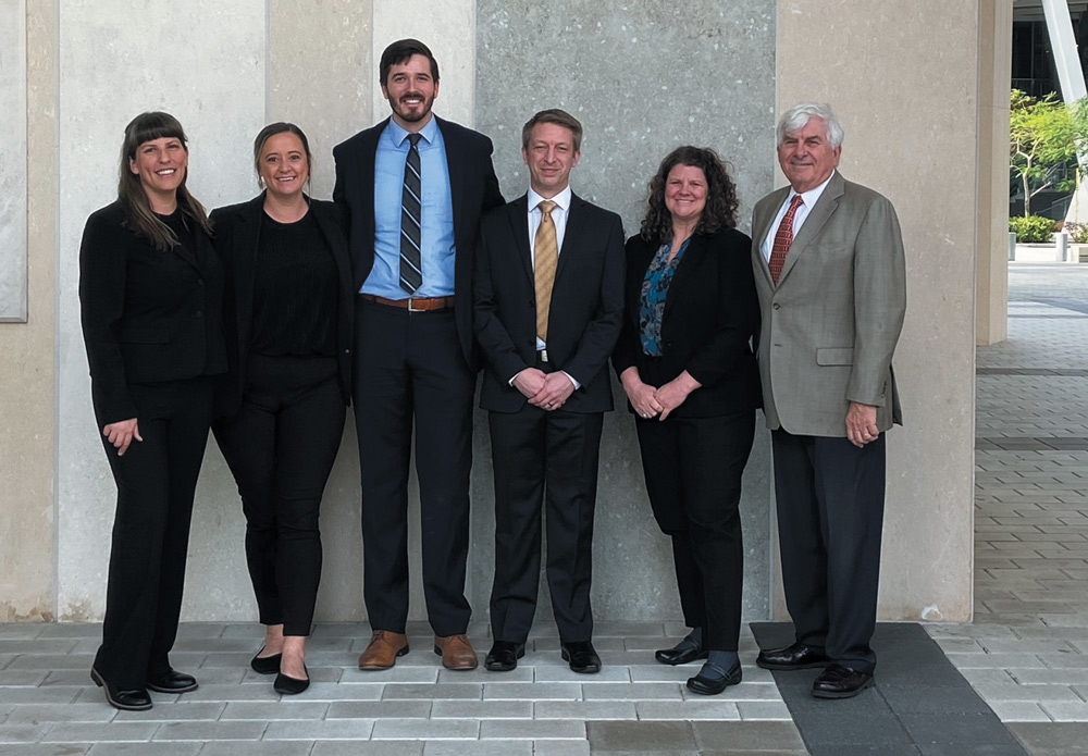 A group of six people in business attire standing in an outdoor setting with stone walls and geometric tile patterns.