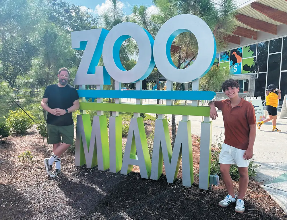 Two people stand by a large "ZOO MIAMI" sign with trees and a building in the background.