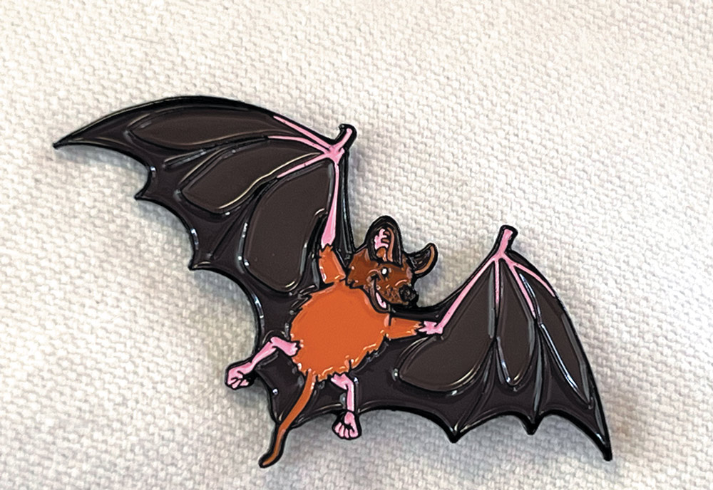 An enamel pin of a bat with an orange body and dark brown wings on a textured gray background.