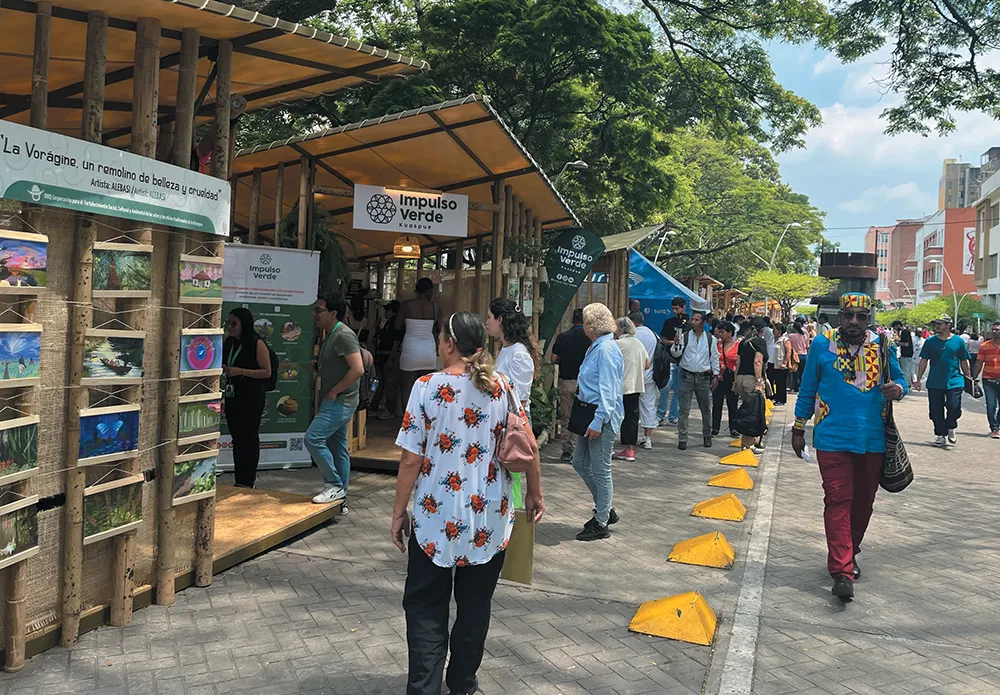 An outdoor market scene with art displays, a booth labeled "Impulso Verde," and several people walking along a pathway with yellow markers.
