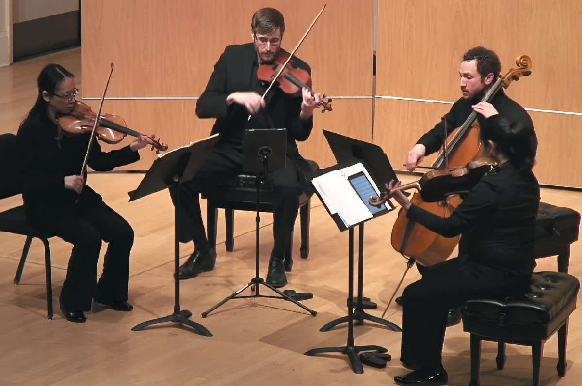 A string quartet performs on a wooden stage, with two violinists, a cellist, and a violist playing their instruments.