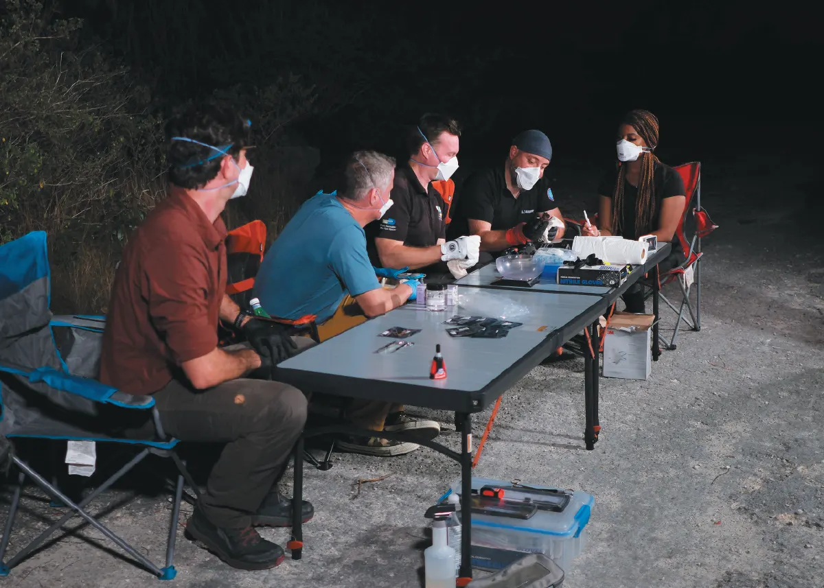 A group of five researchers wearing face masks are seated around a table outdoors at night, examining equipment.