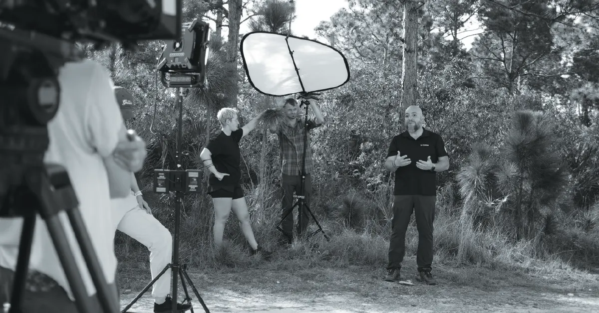 A black and white outdoor video shoot in a wooded area, with a man speaking to the camera while others manage lighting and equipment.