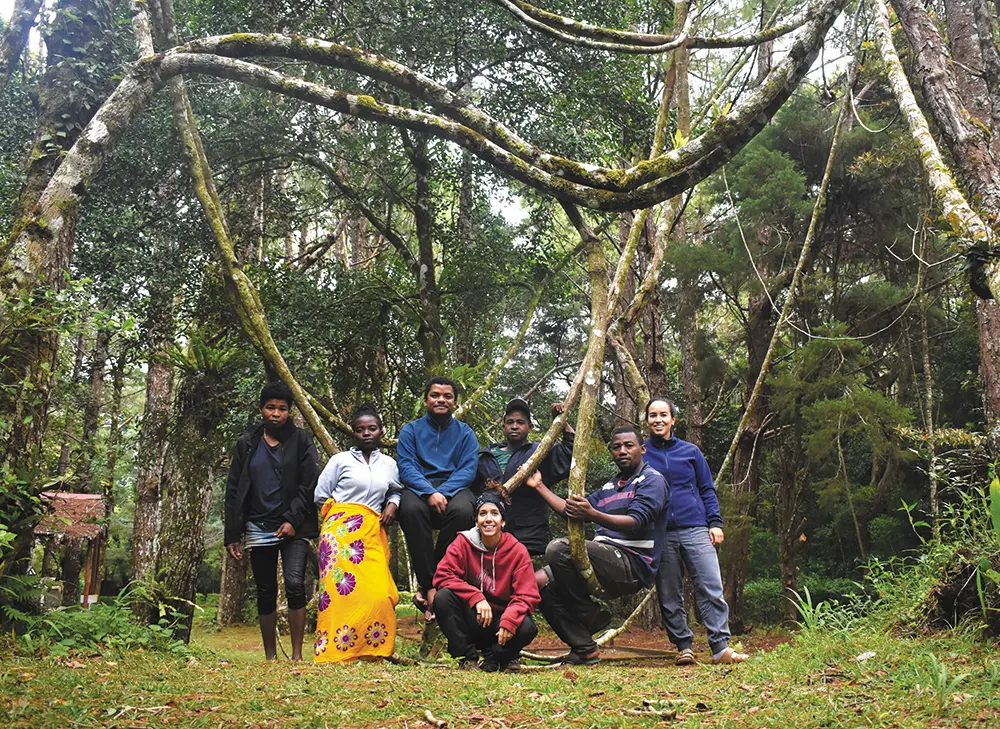 Portrait orientation outdoor photograph of a group of seven young adults that are made up of men plus women who are posing for a photo outdoors in a lush, green forest; Some are standing, others are sitting or crouching, and one man is leaning against a thick vine; They are all smiling and appear to be enjoying the natural surroundings; The individuals pictured here happen to be Carme Tuneu-Corral’s team during a fieldwork expedition in northern Madagascar