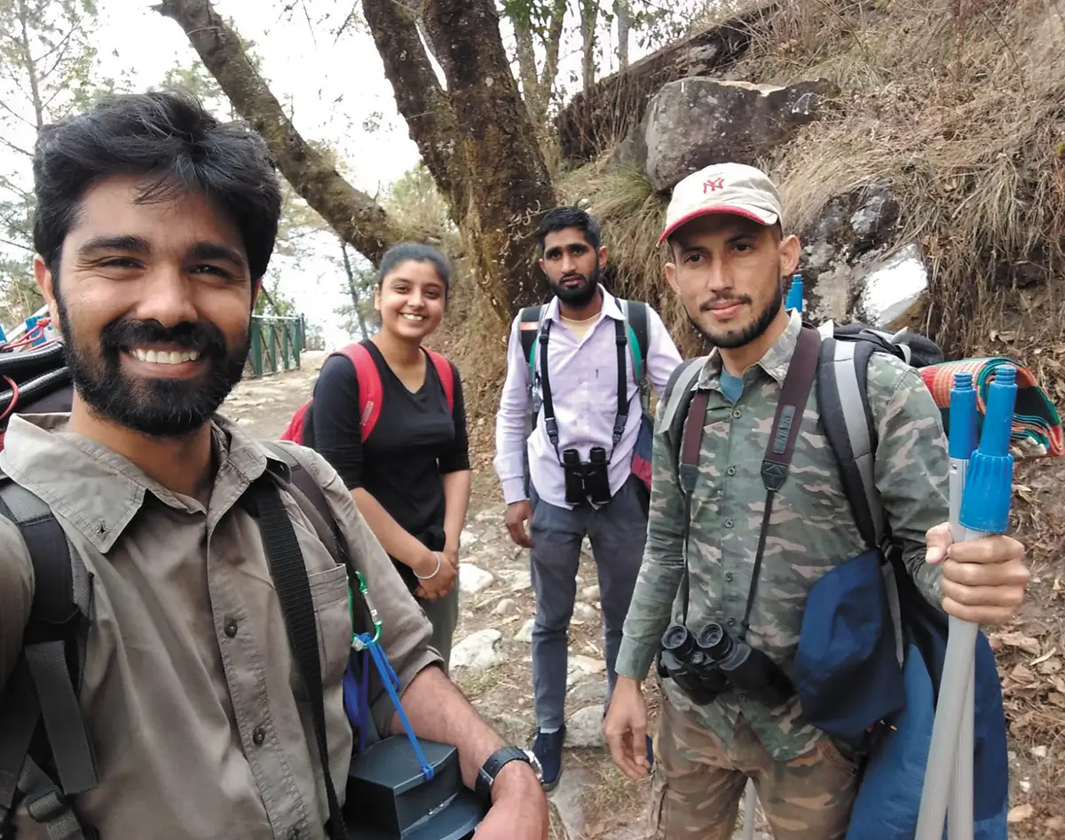 A group of four smiling people, three men and one woman, are posing for a selfie on a rocky, wooded path. They are dressed in hiking or research gear, including backpacks and binoculars. The man on the far left, who is taking the photo, has a beard and is wearing a green shirt. The person on the far right is holding blue poles.