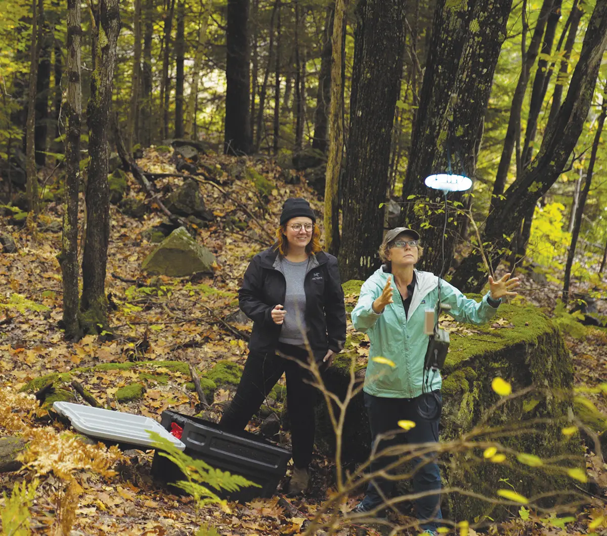 Two women, one in a beanie and black jacket and the other in a light blue jacket and baseball cap, are in a forest. The woman in the blue jacket is looking up and gesturing with her hands toward a small, hovering drone with a glowing light. A black and gray case is on the ground near them. The forest floor is covered in fallen leaves and rocks.