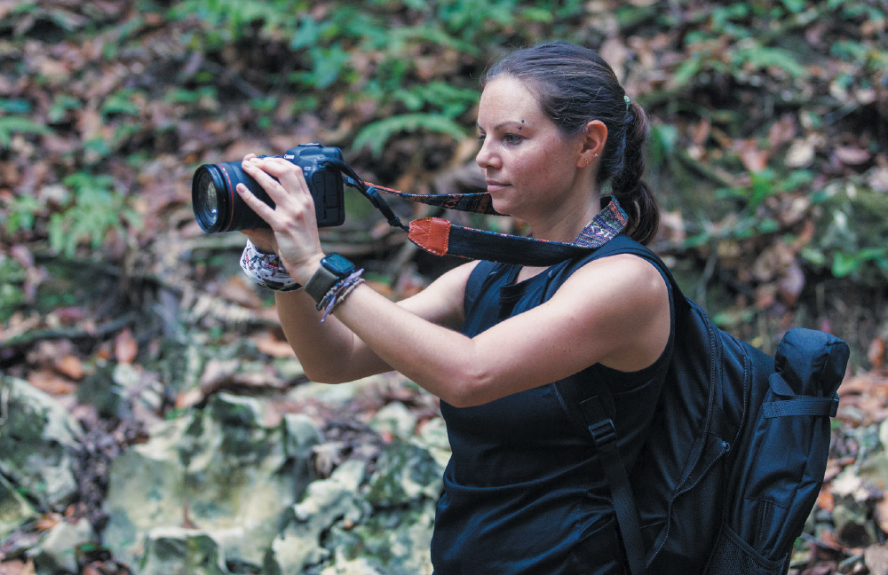 A woman with her hair in a ponytail and a backpack on is holding a camera with a large lens. She is wearing a black tank top and a watch. She is standing outdoors in a forest and appears to be taking a photo of something out of frame to the right.