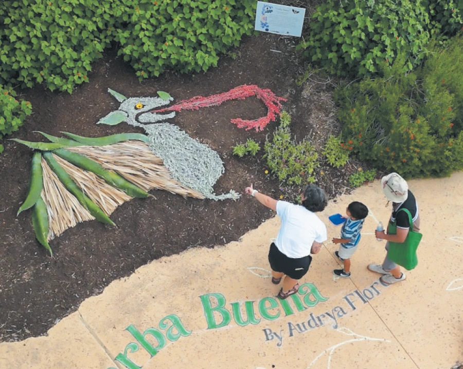 A high-angle shot shows a family of three — a woman, a man, and a small child — walking along a paved path. To the left of the path, a striking land art installation depicts a bat crafted from various natural materials. The adults and the child are all looking at the bat, with the woman pointing towards it.