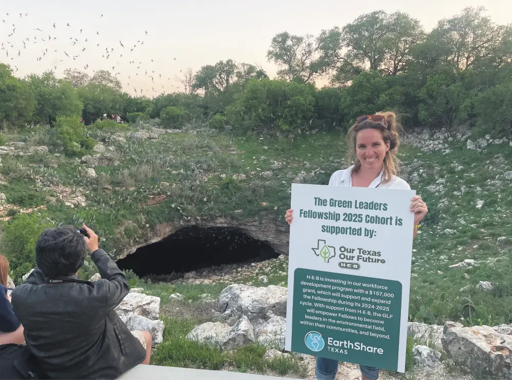Outdoor photograph close-up view of Emily Hutcheson, a smiling woman holding a sign that reads, The Green Leaders Fellowship 2025 Cohort is supported by: Our Texas Our Future H-E-B, EarthShare Texas; The sign mentions a $157,000 grant for workforce development; She is standing outdoors in a hilly, green landscape area filled with green lush trees, boulder rocks, and a large cave entrance behind her at Bracken Cave; A slight partial cropped view of a random man and a few other individuals are situated seated down on a concrete bench to the left of Emily as the random man is taking a photo of the Bracken Cave's topography outdoor nature surroundings; In the sky, a large swarm of bats is visible, flying out of the cave