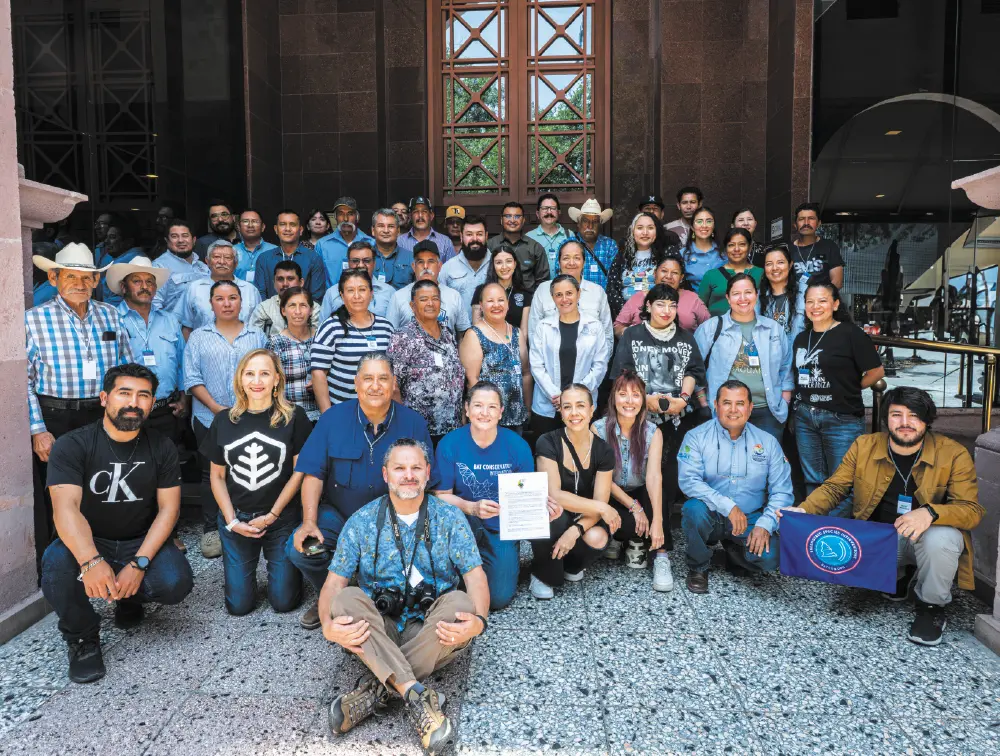 Photograph of a large group of approximately 40 to 50 diverse people who are gathered for a group picture outdoors in front of a stone building with large windows; Many people are standing in the back, while a smaller group is kneeling or crouching in the front; One woman in the center is holding a framed document; All these individuals pictured here happen to be BCI and partners gathered together to focus on protecting agave for bats