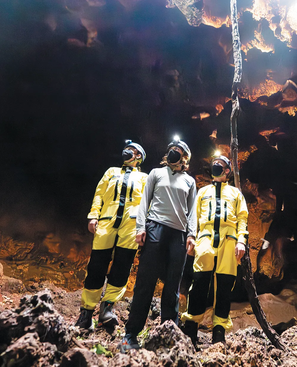 Three researchers in a dark cave wear protective suits, face masks, and headlamps as they look upward toward a light source near a rocky formation.