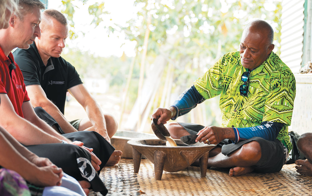 A man in a bright green patterned shirt performs a traditional kava ceremony, pouring liquid from a coconut shell into a wooden bowl as two men watch respectfully.