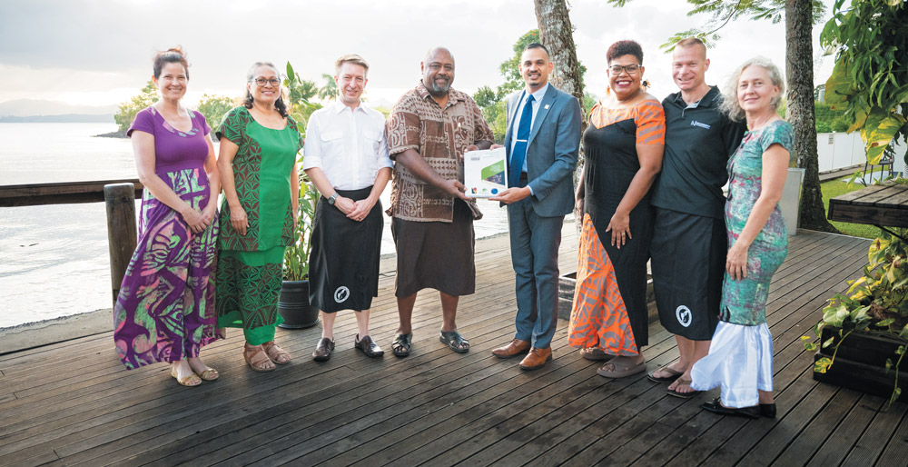 A diverse group of eight people stand in a row on a wooden deck overlooking the water, with two men in the center holding an official conservation document together.