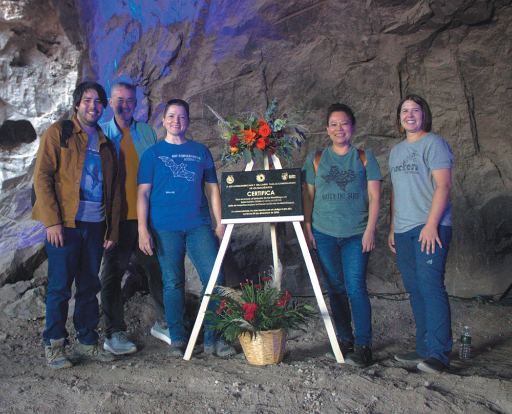 A group of five people stand inside a cave next to a formal commemorative plaque on an easel. The plaque is decorated with red and orange flowers.