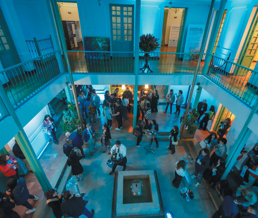 A high-angle view of a social gathering in a multi-story indoor courtyard. People are mingling and talking near a central fountain, with the space bathed in blue atmospheric lighting.