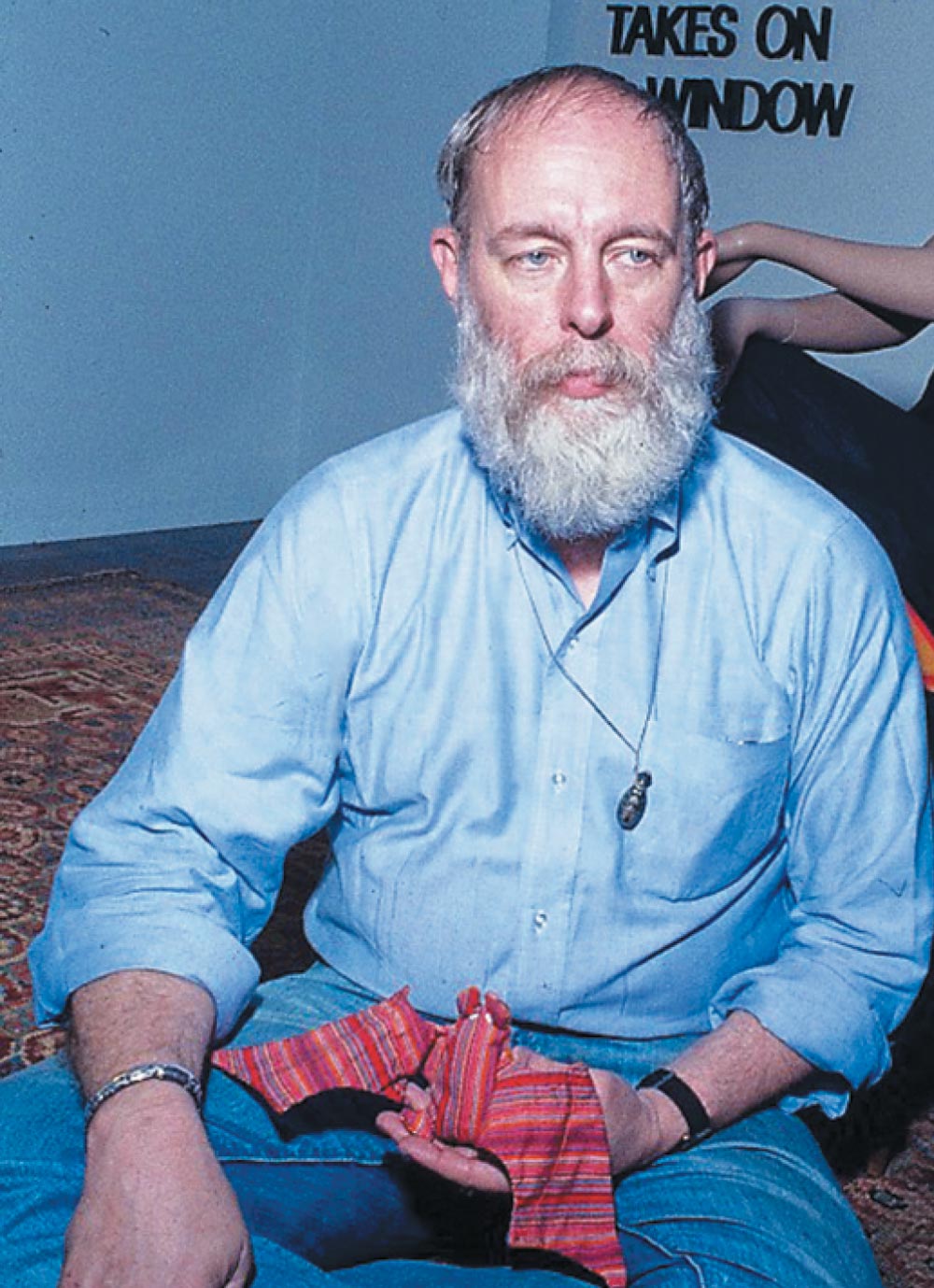 A bearded man holding a striped fabric, sitting on the floor, against a blue wall background.