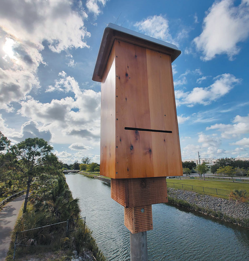 A wooden bat house on a pole near a waterway under a partly cloudy sky.