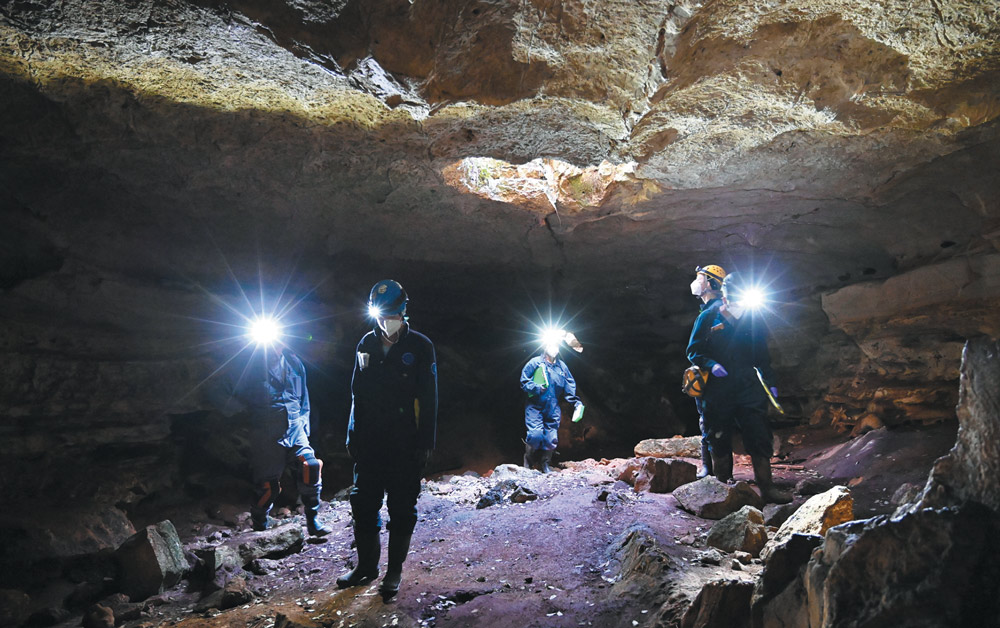 Five researchers wearing jumpsuits, masks, and bright headlamps explore a large, dark underground cave with a rocky ceiling and a small natural opening above.