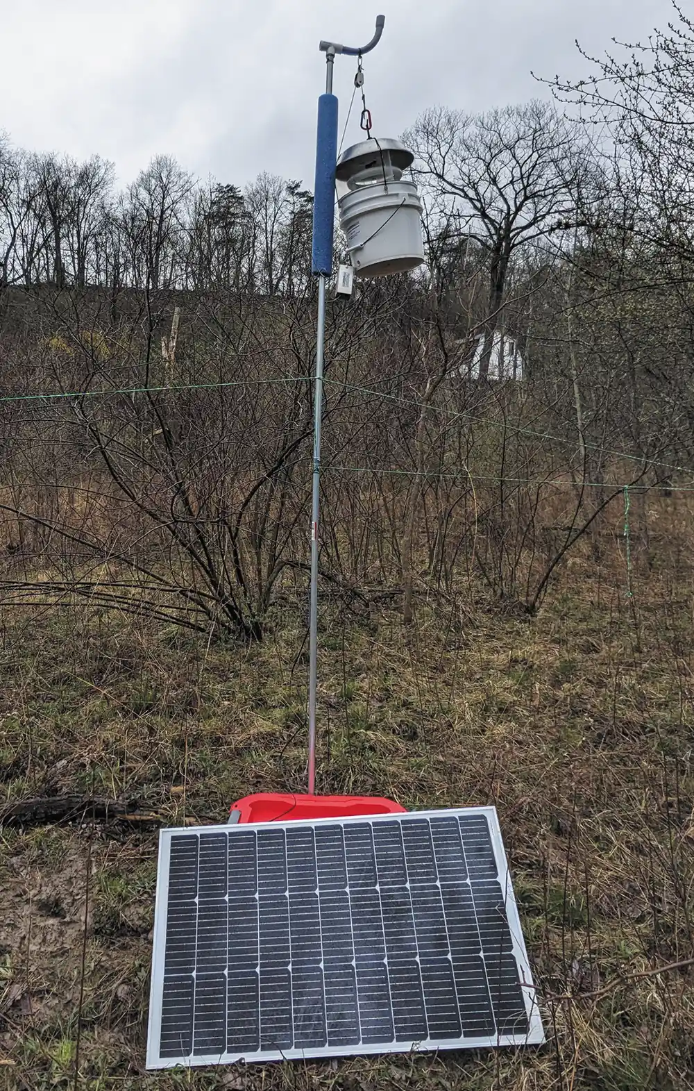 A scientific monitoring station in a field features a solar panel on the ground connected to a tall metal pole holding a white bucket-like device and foam insulation.