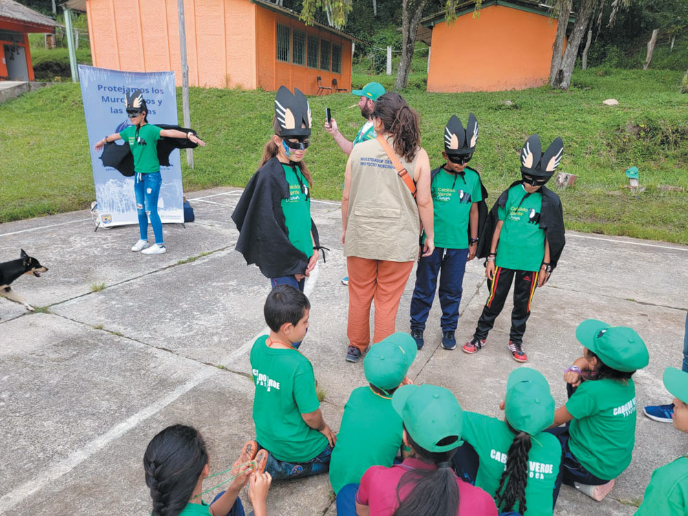 A group of children in green shirts and bat-themed masks and capes participate in an outdoor educational activity led by an adult researcher in a tan vest.