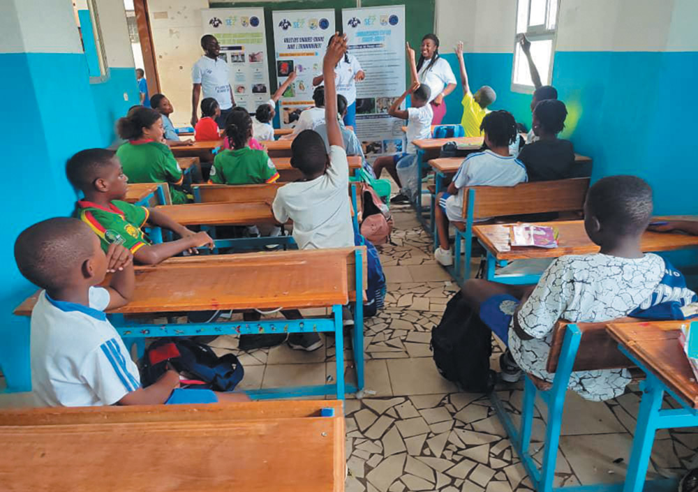 A classroom full of students with their hands raised, participating in an educational session led by three instructors standing in front of informative banners and posters.