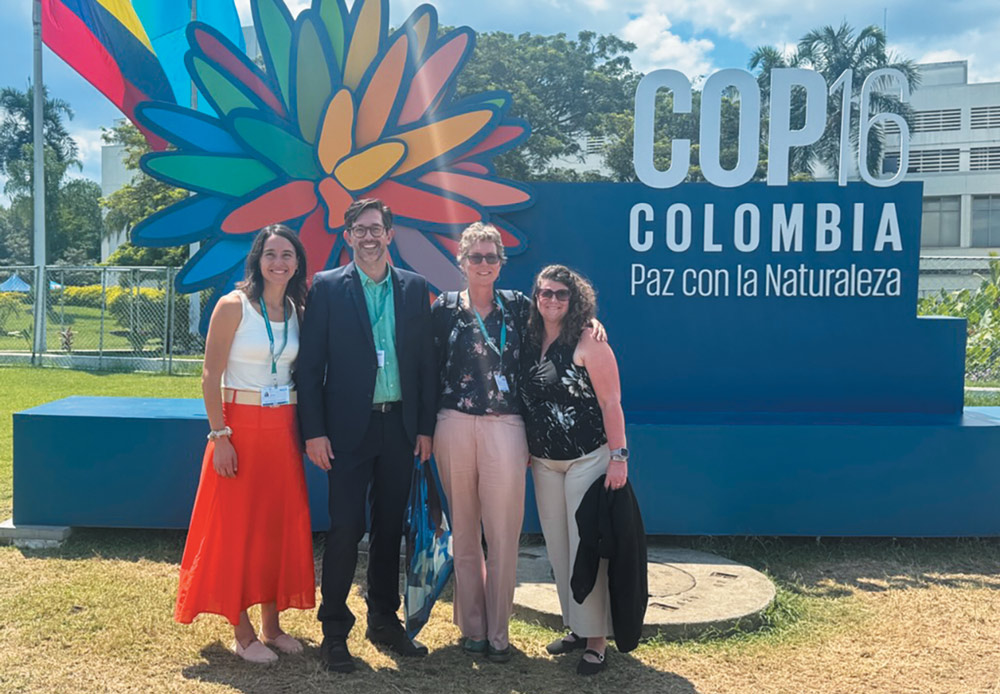 Four professionals pose for a photo in front of a large blue "COP16 COLOMBIA" sign featuring a colorful flower logo and the slogan "Paz con la Naturaleza" under a bright sky.