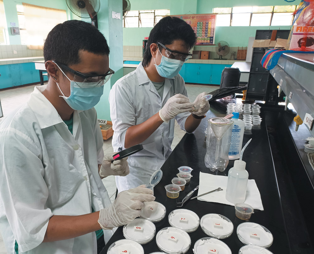 Two researchers in white lab coats, face masks, and gloves carefully examine biological samples in petri dishes using a magnifying glass in a bright, organized laboratory.