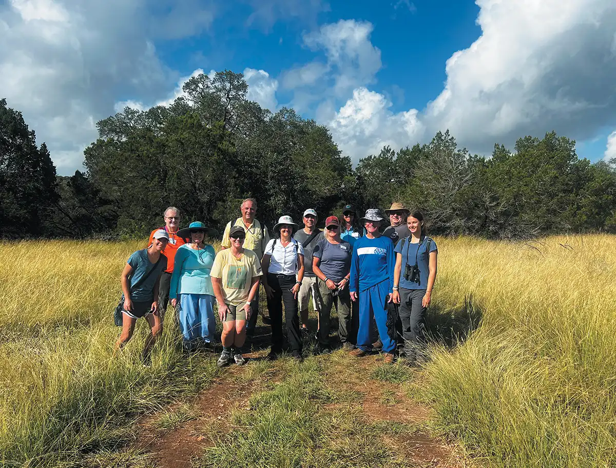 A large group of people in hiking attire pose for a photo in a golden grassy field. A line of dark green trees and a bright, cloudy sky form the backdrop.