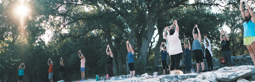 A group of people practice yoga in a sunlit outdoor area. They stand in a row with arms raised toward the sky, surrounded by large, leafy oak trees.