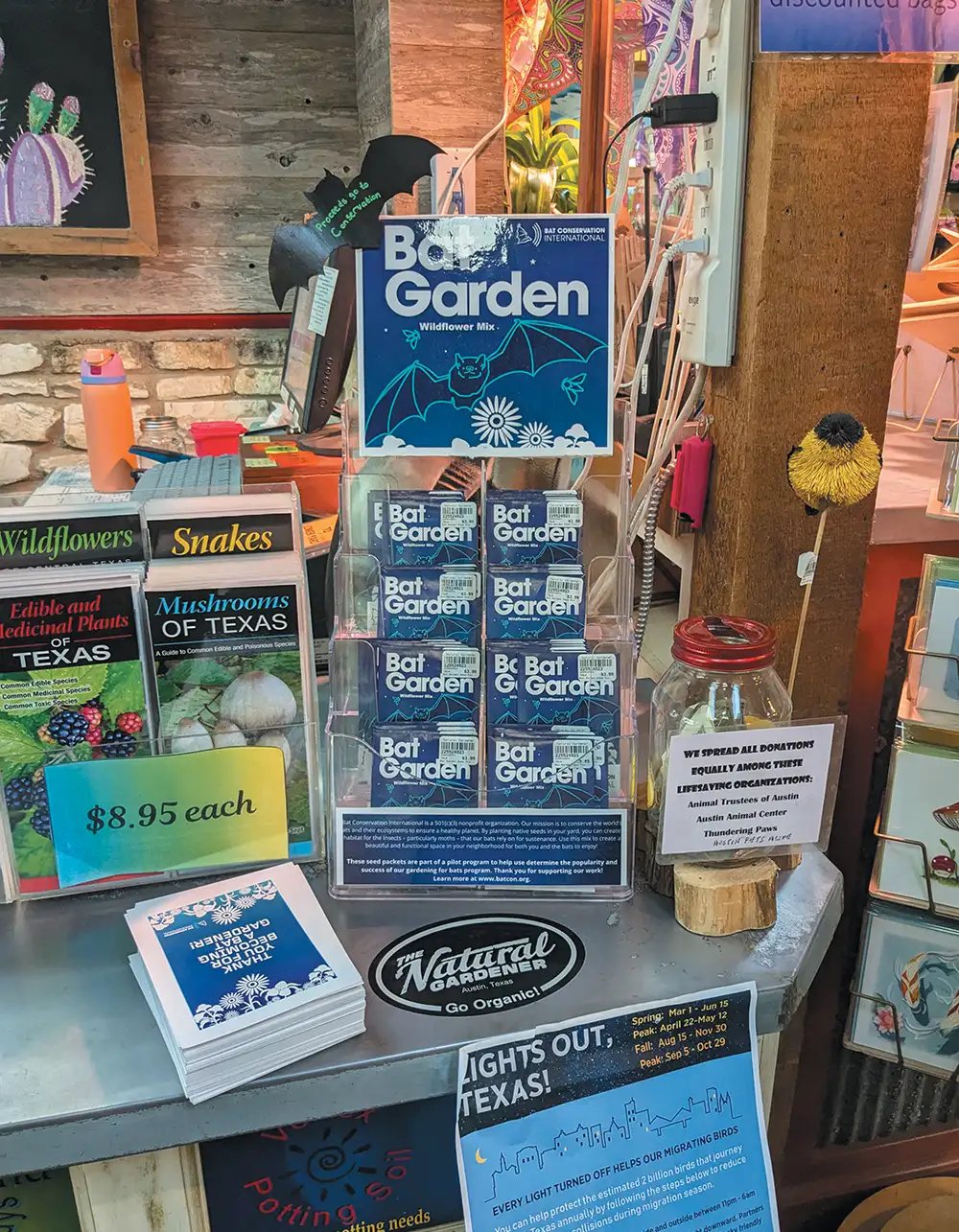 A retail display features "Bat Garden" wildflower seed packets. Surrounding items include nature books, a donation jar, and "Lights Out, Texas!" flyers.