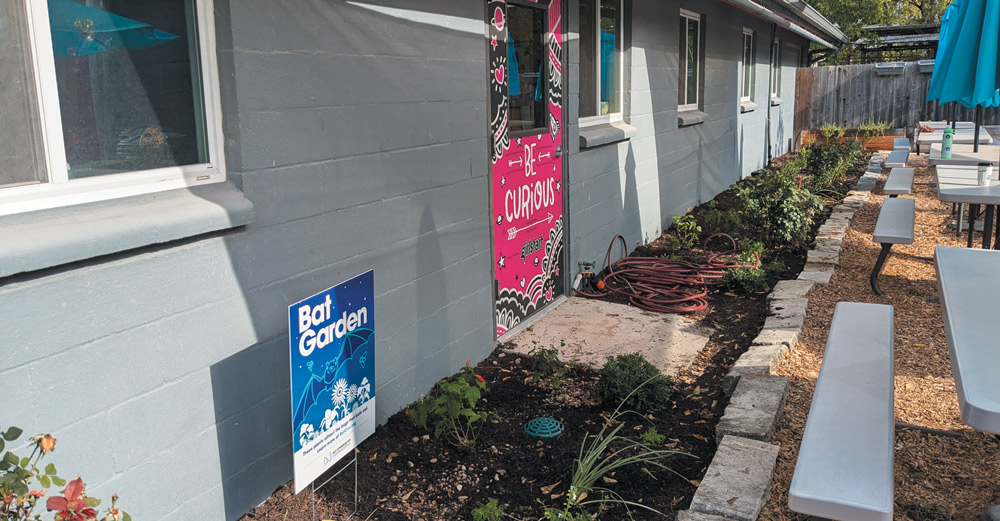 A "Bat Garden" sign stands in a freshly mulched garden bed alongside a gray building. A pink door with "BE CURIOUS" text and outdoor seating are visible.