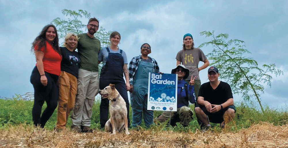 A diverse group of seven people and a dog pose behind a "Bat Garden" sign in a grassy outdoor area under an overcast, cloudy sky.
