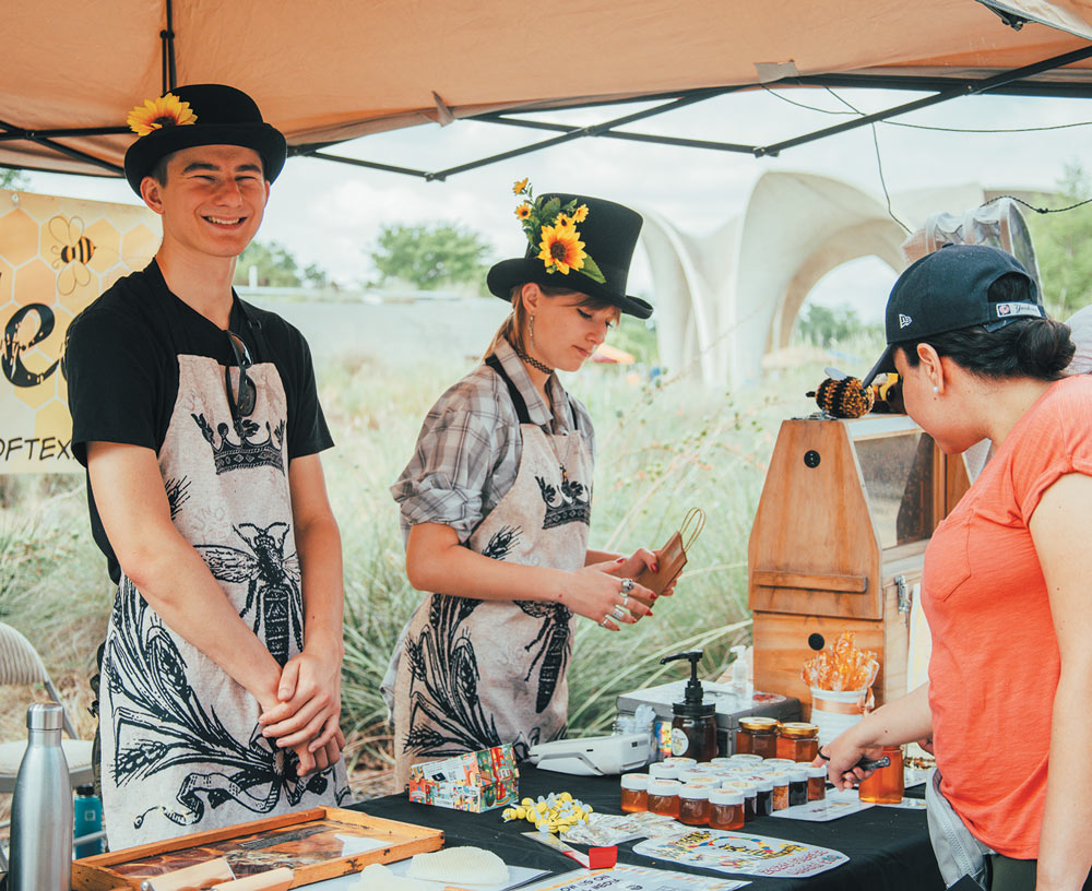Two vendors in bee-themed aprons and hats decorated with sunflowers serve a customer at an outdoor market stall featuring honey and nature products.