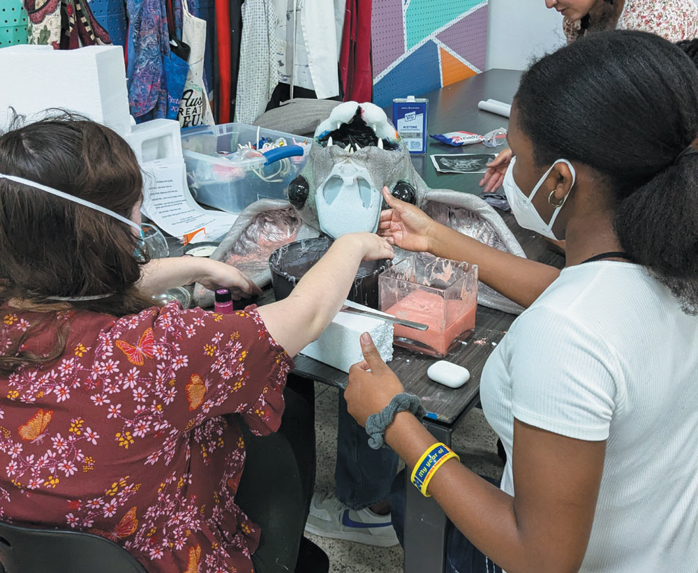 Two people wearing face masks work on a large, detailed bat sculpture at a table, applying materials to the head and wing sections.