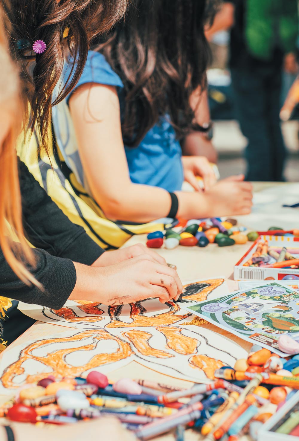 Close-up of children's hands using colorful crayons to fill in large bat-themed drawings at a crowded outdoor activity table.