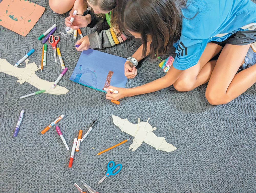 An overhead view of two children sitting on a carpeted floor, using markers and scissors to create bat-themed crafts and drawings.