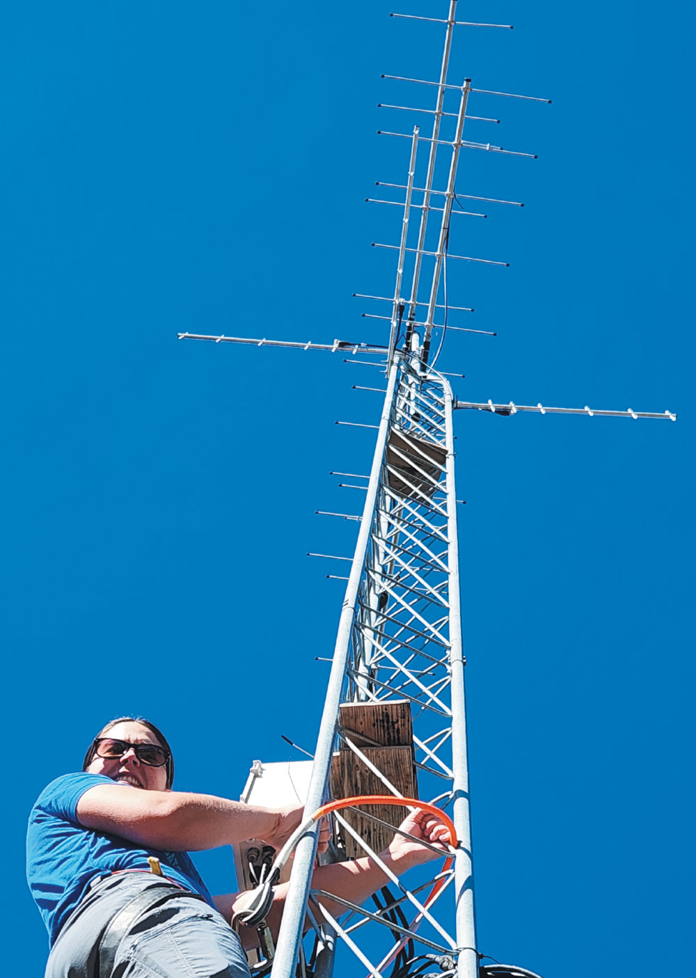 A person wearing sunglasses and climbing gear works on a tall, silver lattice telemetry tower. The metal tower is topped with multiple antenna arrays against a clear, bright blue sky.